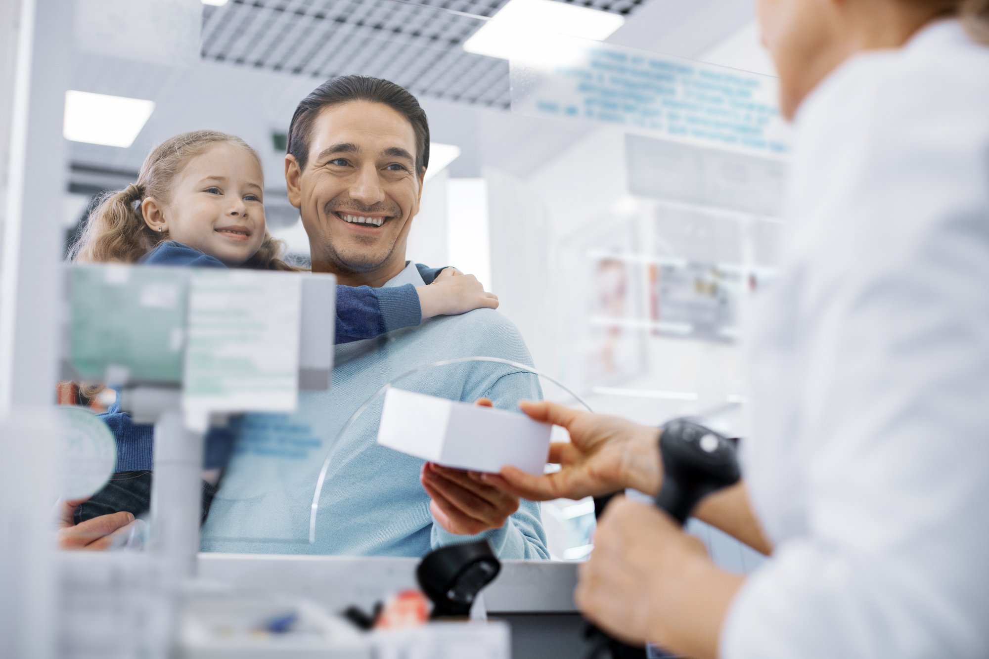 Père souriant tenant un jeune enfant tout en recevant un colis d'un pharmacien au comptoir d'une pharmacie.