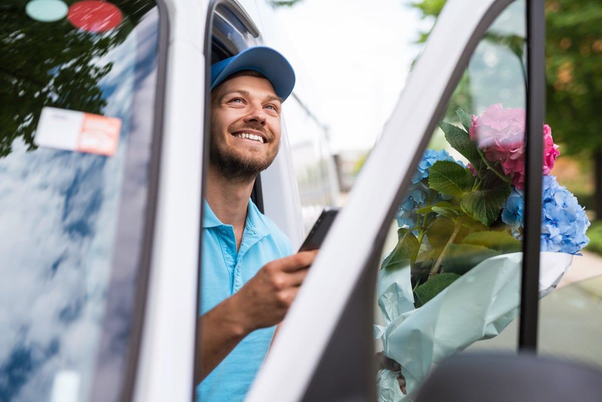 Livreur souriant tenant un smartphone dans une camionnette avec un bouquet de fleurs, suggérant une livraison florale.